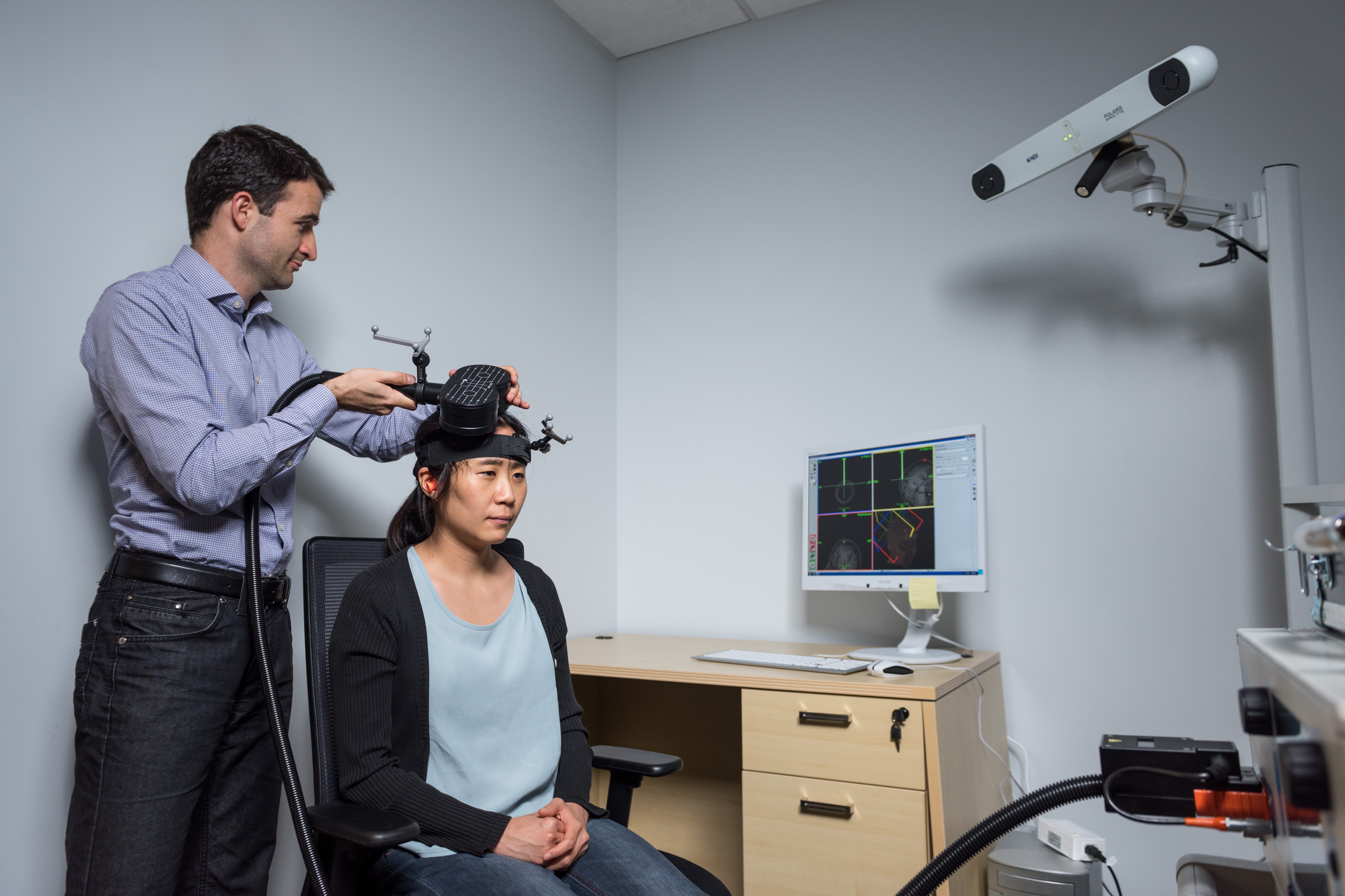 Researcher adjusting a device on another person’s head in a lab, with a computer displaying brain imaging data and a mounted camera in the background.