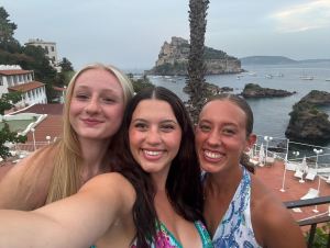 Students from Psychology 3000, including Abby Davis, Bailey Benak, and Sarah Peach took their studies global this summer, exploring how culture shapes stress and well-being — one breathtaking view at a time. Three girls on a clifftop overlooking blue water taking a selfie.