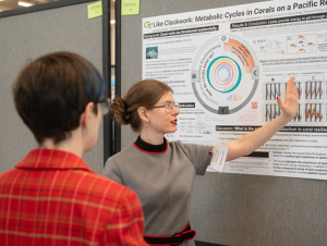 A man looks at a woman who is explaining her research via a poster.
